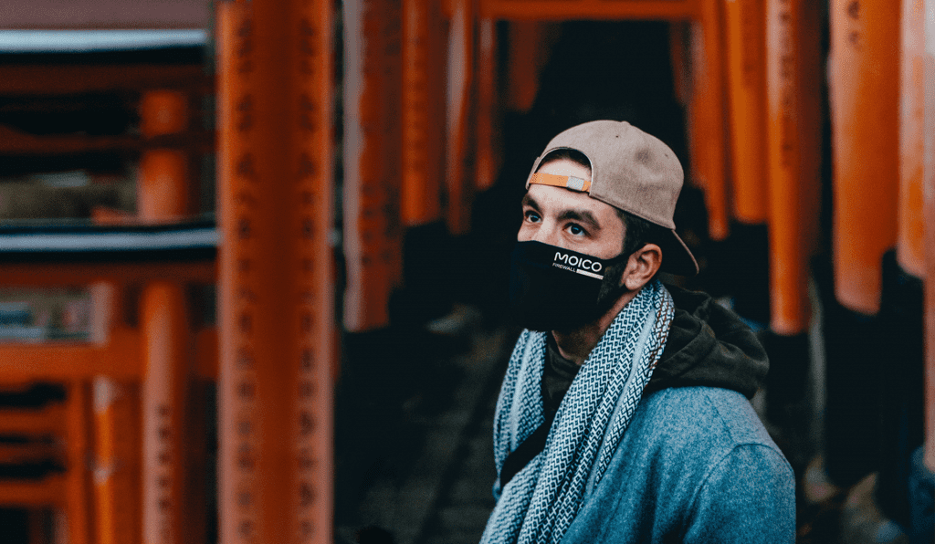 Na zdjęciu widzimy mężczyznę stojącego na tle jakiś pomarańczowych, metalowych regałów. Mężczyzna nosi jasną czapkę, niebieską kurtkę, szalik i czarna maskę zakrywającą nos i usta z napisem MOICO.