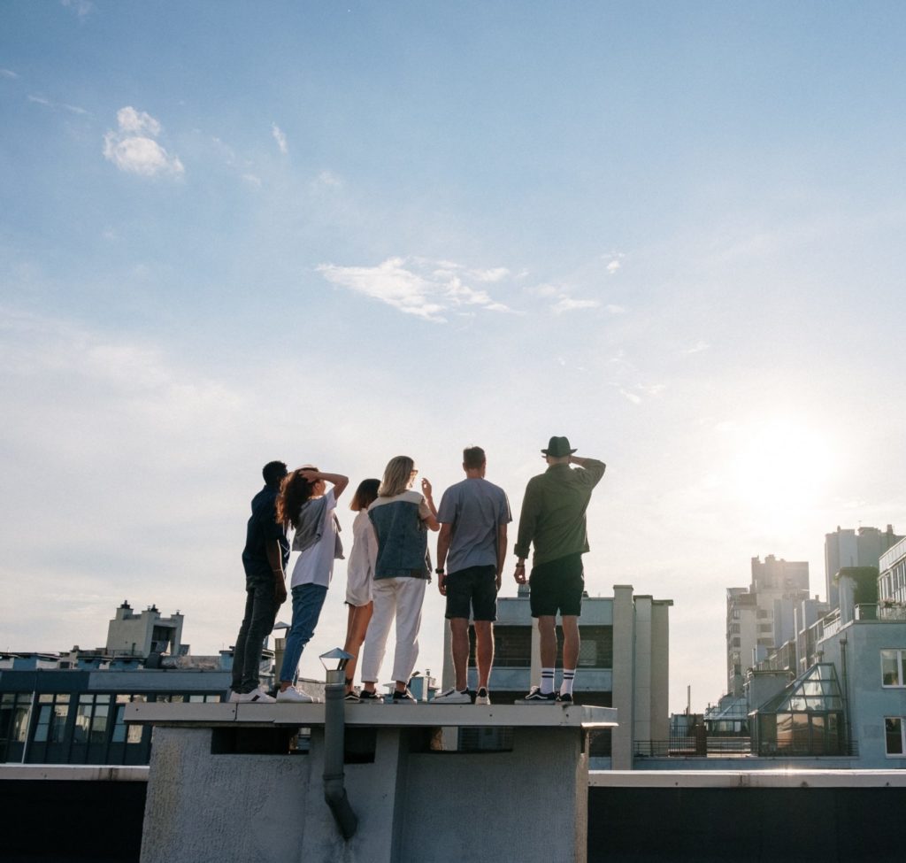 Obraz przedstawia grupę sześciu osób stojących na dachu i patrzących na horyzont. Są ubrani swobodnie, a sceneria to jasne niebo z kilkoma chmurami i promieniami słońca. W tle widać miejską panoramę z budynkami, co dodaje nowoczesnego klimatu.