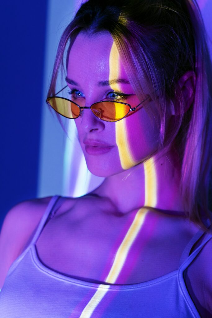 Na obrazku widać kobietę w okularach, które są oparte na nosie. Kobieta ma na sobie koszulkę na ramiączkach, z kolorową projekcją świetlną na górnej części ciała. Projekcja świetlna składa się z odcieni fioletu i żółtego, tworząc interesujący efekt wizualny.