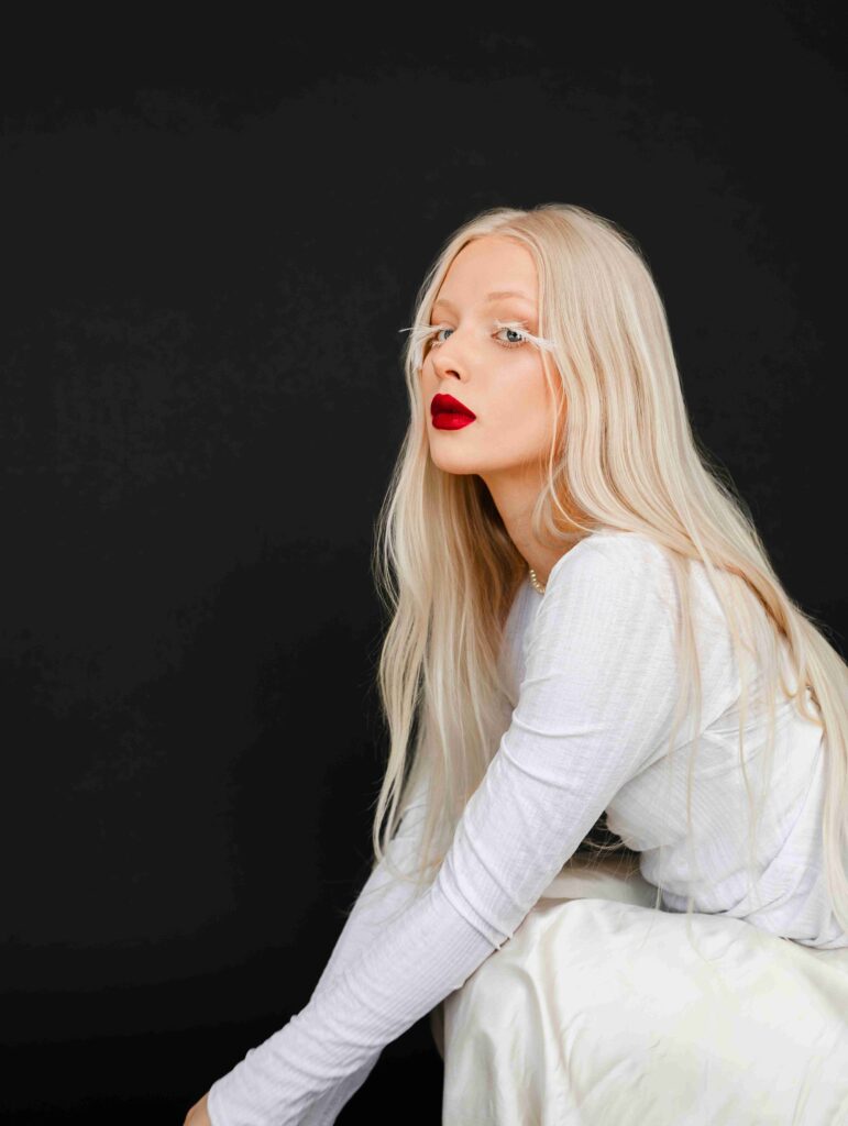 a jednolitym czarnym tle siedzi kobieta o blond, prostych włosach, ubrana w białą bluzkę i białą spódnicę. Jej czerwone usta kontrastują z jasną cerą i stonowaną stylizacją. Spojrzenie kobiety skierowane lekko w bok.